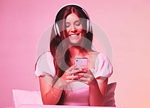 Pink, light and music by woman in a bed with headphones, social media and streaming on wall background. Podcast, texting
