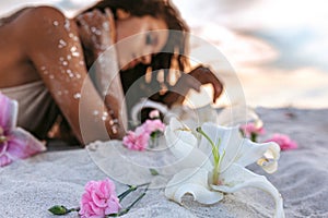 Pink lilies lying on sand foreground and young beatiful woman lying on sand at background on the beach at sunset