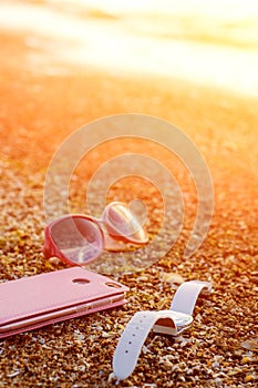 Pink Mobile Phone, White Wrist Watch And Pink Glasses On The Sandy Sea Beach With Waves And Sunlight. Modern Smart Phone
