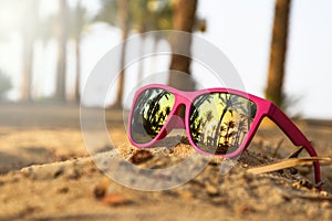 Pink modern sunglasses on the beach sand with palm trees and sunset