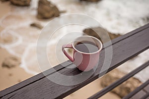 Pink mug of black coffee on rail above ocean shore