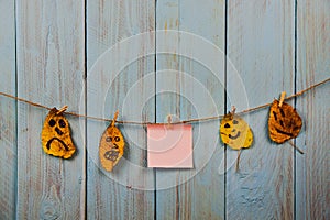 Pink note papers, and funny faces on the autumn leaves hanging on the rope against blue rustic wooden background. Copyspace.