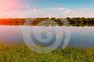 Pink orange summer evening sky. Sun rays and clouds reflecting in water. Meadow and forest on banks