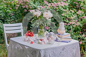 Pink peonies in a glass vase and books in lilac covers, sweets, marshmallows and ripe strawberries in a bowl on a white table