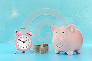 A pink pig piggy bank on a blue background. Nearby is a small pink fridge and three pile of coins.