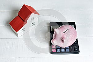 Pink pig piggy bank, calculator and breadboard model of a house with a red roof on a light background. Concept of renting, buying