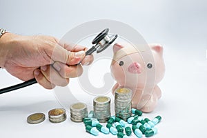 A pink piggy bank with a bandage at head being placed stethoscope at head with stacking coins and blue green capsules pills