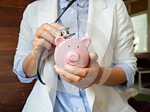 Pink piggy bank examined by a female doctor. Using stethoscope. Wooden background for copy space. Financial Health concept