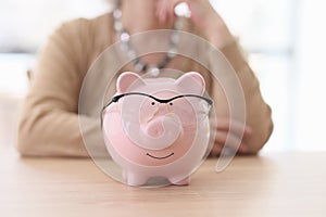 Pink piggy bank with glasses stands on wooden table at lady