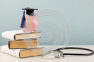 Pink piggy bank with graduation cap on stack of books and stethoscope. Concept of the costs of medical education