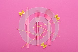 Pink plastic knife, fork and spoon, spaghetti and uncooked farfalle pasta isolated on pink.