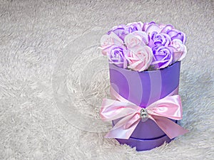 Pink and purple roses in a cylindrical purple box tied with a pink silk ribbon on a white fur background.