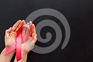 Pink ribbon in hands as symbol of breast cancer awareness on black background top view copy space