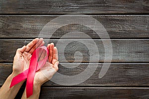 Pink ribbon in hands as symbol of breast cancer awareness on dark wooden background top view space for text