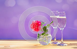 Pink rose and two glasses of champagne on purple background. Greeting card with space for your text