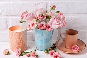 Pink roses flowers in blue cup, burning candle and little cup for coffee against white brick wall.