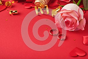 Pink roses flowers, wedding rings, champagne, gift, golden ribbons and confetti hearts on red background. Top view with space for