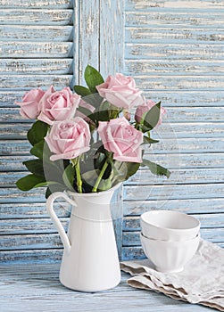 Pink roses in a white enameled pitcher and ceramic white bowls on blue wooden rustic background. Kitchen still life