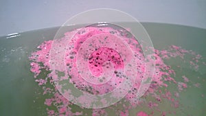 Pink round bath bomb dissolves in water. In the frame edge of the white bath photo