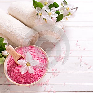 Pink sea salt in bowl, towels and flowers on white wooden backg