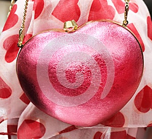 Pink sparkly heart purse with a gold chain strap hanging in front of a red heart print scarf