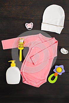 Pink summer bodysuit, hat, two sylicone pacifiers and two wooden and plastic rattles on brown wooden background with copy space.