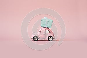 Pink toy retro model car delivering gift box for Valentine`s day on pink background. Volkswagen Beetle on pink background.