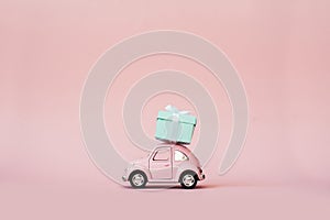 Pink toy retro model car delivering gift box for Valentine`s day on pink background. Volkswagen Beetle on pink background.