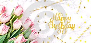 Pink tulips and gold Happy birthday letters with festive straws on white marble background. Spring and celebration