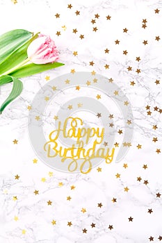 Pink tulips and gold Happy birthday letters on white marble background with golden confetti. Spring and celebration