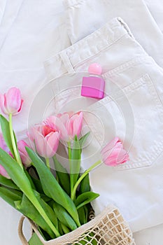 Pink tulips in macrame bag and perfume bottle on white jeans background