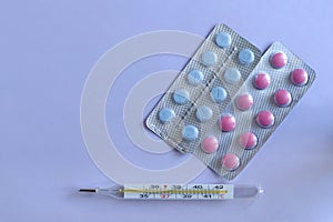 Pink, white and blue pills in blisters and a thermometer with copy space