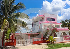 Pink and white Mexican three story house with red fence and palm and flowering trees against a beautiful blue sky with fluffy clou