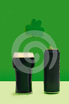 A pint of black stout beer glass and beer can standing alone in front of green clover above. Lucky pub minimal concept