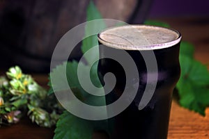 Pint of dark beer foam beautifully on a barrel with a leaf of hops background