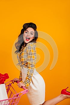 Pinup girl posing with shopping cart. Amazed brunette woman having fun on yellow background