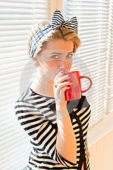 Pinup girl with red lips nails drinking coffee or tea holding red cup & looking at camera over white sun blinds