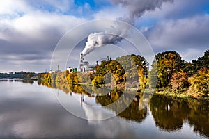 Pipes of woodworking enterprise plant sawmill near river with autumn red yellow trees. Air pollution concept. Industrial landscape