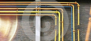 Pipes in yellow, orange and blue bend at right angles in front of a brick wall, with light beams