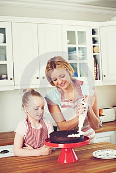 Piping on the final finishings. Cute little girl icing a cake with her mom in the kitchen.