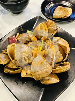 Pipis seafood in black plate, one isolated in black round sauce plate, on white table.