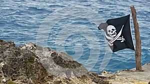 Pirate black flag Jolly Roger with painted white skull and two crossed pirate sabers stands on rocky shore with blue sea