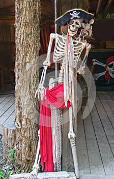 Pirate skeleton with peg leg and eye patch and hat and dead parrot on shoulder propped on tree trunk pillar with pirate flag in