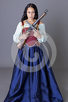 A pirate woman wearing a brocade corset and silk skirt and  holding guns against a grey backdrop