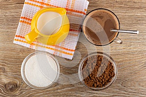 Pitcher with milk on napkin, bowls with cocoa powder and sugar, cocoa with milk, spoon in cup on table. Top view