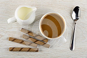 Pitcher with milk, wafer rolls with chocolate filling, coffee with milk in cup, spoon on table. Top view