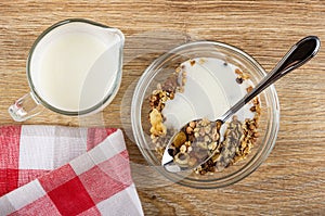 Pitcher with yogurt, spoon in bowl of granola with banana and chocolate with yogurt, napkin on table. Top view