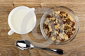 Pitcher with yogurt, transparent bowl of granola with banana and chocolate, spoon on wooden table. Top view