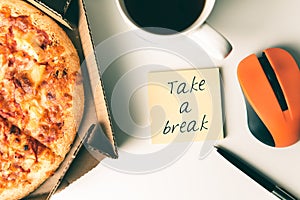 Pizza in box, cup of coffee, laptop, computer mouse, pen and sticker with text TAKE A BREAK on table in office.
