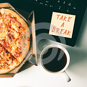 Pizza in box, cup of coffee, laptop and sticker with text TAKE A BREAK on table in office.